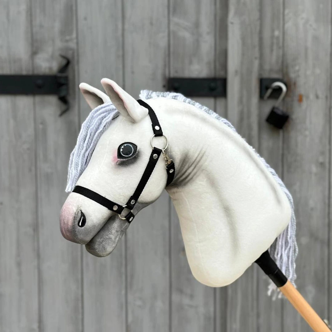 The Hobby Horse Atlas with Black Halter by Hobby Horse LarDen, featuring a white Appaloosa design and gray yarn mane, stands before a wooden door secured with a metal latch and padlock.
