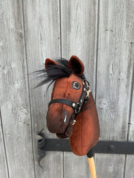 Hobby Horse LarDen’s Benny with Black Halter (size XS) is a brown plush hobby horse inspired by a Belgian Warmblood, featuring a black mane, mounted on a wooden stick and shown in front of a weathered wooden fence.