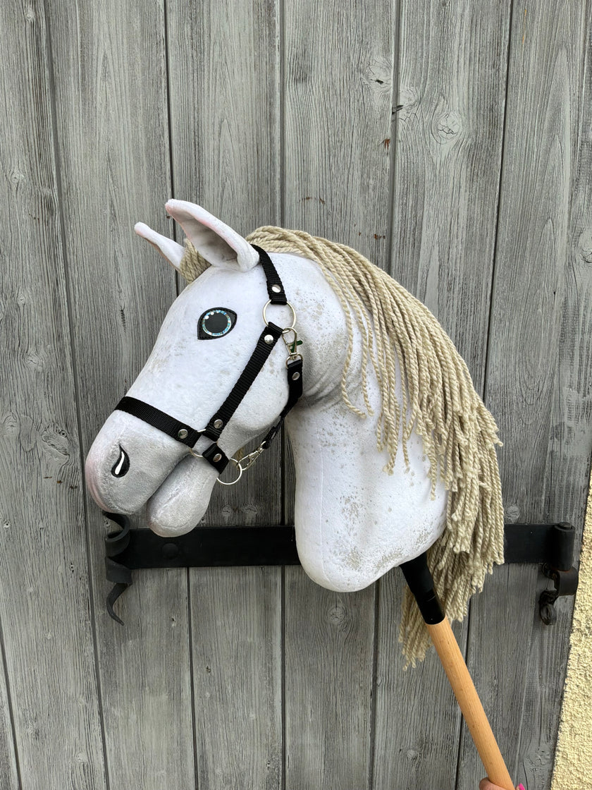 The Hobby Horse Victory with Black Halter (size M) by Hobby Horse LarDen, featuring a white coat and beige yarn mane, rests on its wooden stick against a weathered gray wooden door.