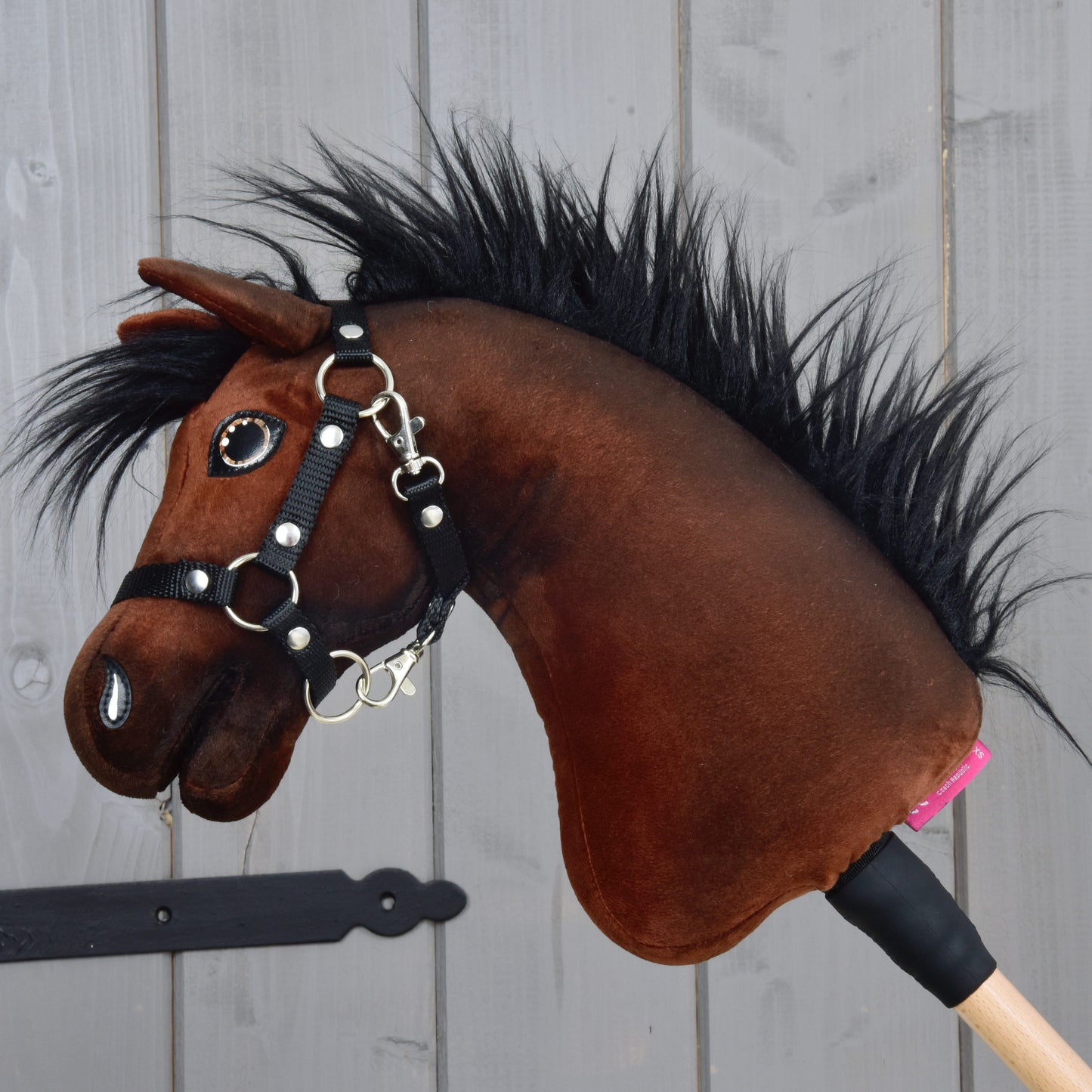 Hobby Horse LarDen’s Hobby Horse Benny with Black Halter (size XS), inspired by a Belgian Warmblood, features a plush brown body, black mane, and halter on a wooden stick, shown against a light gray wooden panel background.