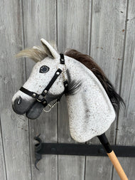 The Hobby Horse LarDen "Hobby Horse Luna with Black Halter" (size S) features a realistic gray Appaloosa design, black and white mane, and expressive stitched details, mounted on a wooden stick before a weathered fence.
