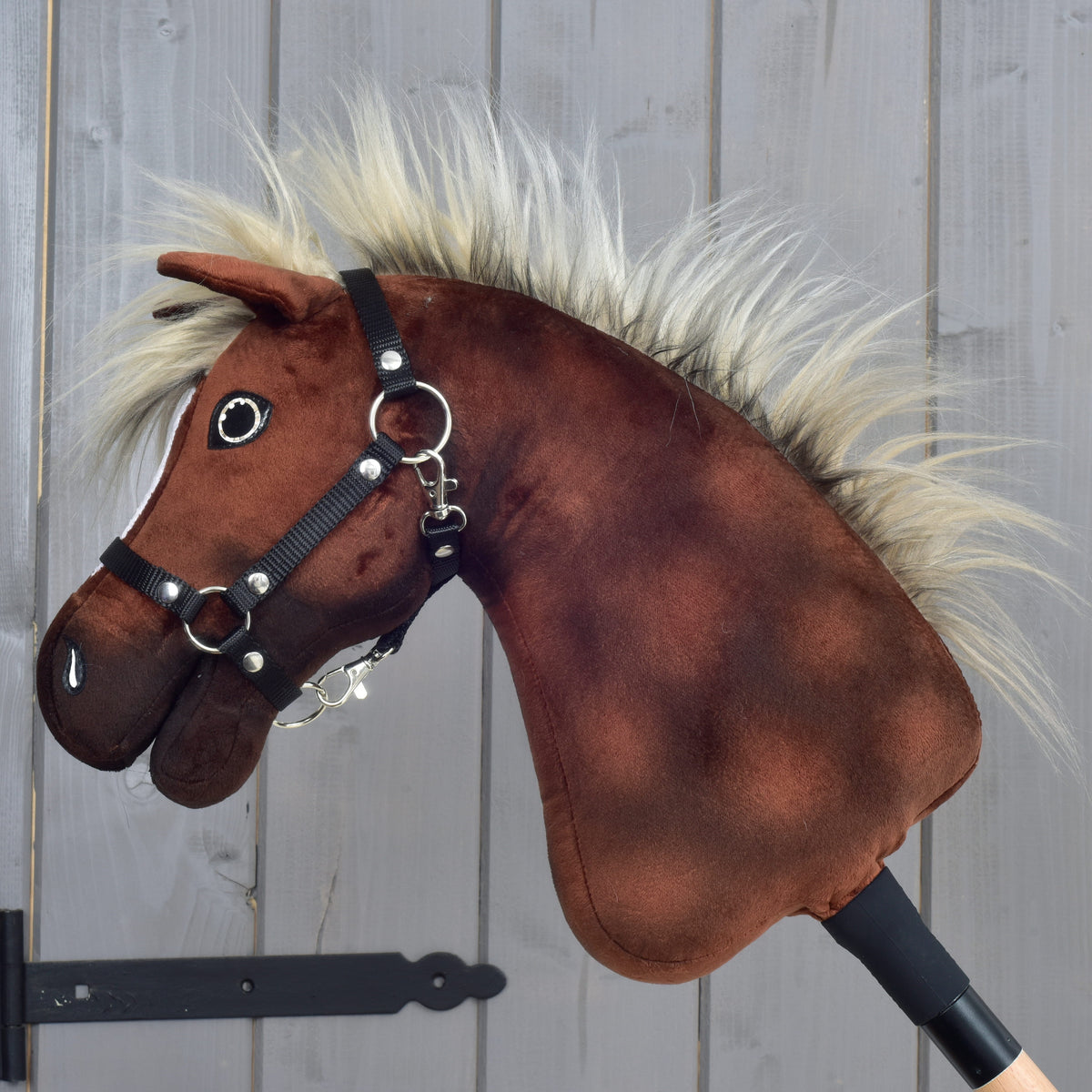 The Hobby Horse LarDen “Hobby Horse Cinnamon with Black Halter - size S” features a soft brown plush head with a bridle and fluffy beige mane, mounted on a stick and reminiscent of an Appaloosa stallion, set against a wooden background.