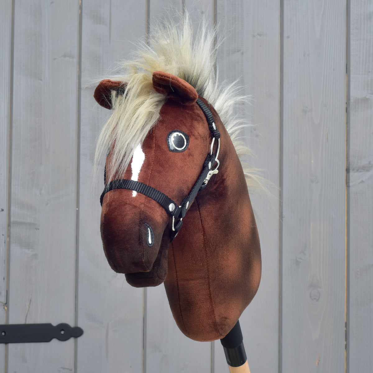 A plush brown “Hobby Horse Cinnamon with Black Halter - size S” by Hobby Horse LarDen, featuring a white blaze, black halter, and fluffy blond mane, is mounted on a wooden stick in front of a gray wooden fence.