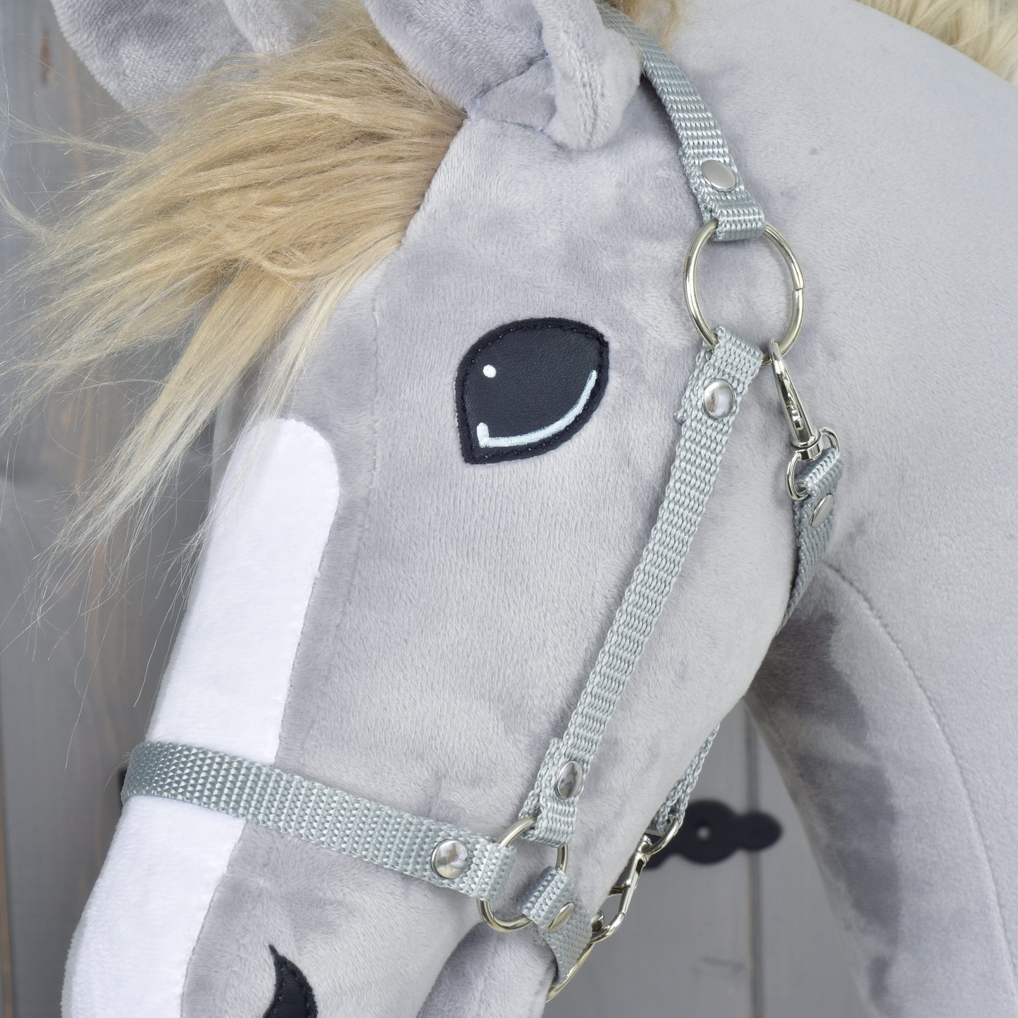A close-up of a gray plush toy horse with a white snout, black stitched eye, and wearing the Set Start Silver (halter + cordeo + lead rope + reins) by Hobby Horse LarDen. Its mane is fluffy and light blonde, making it a charming addition to any collection of hobby horse accessories.