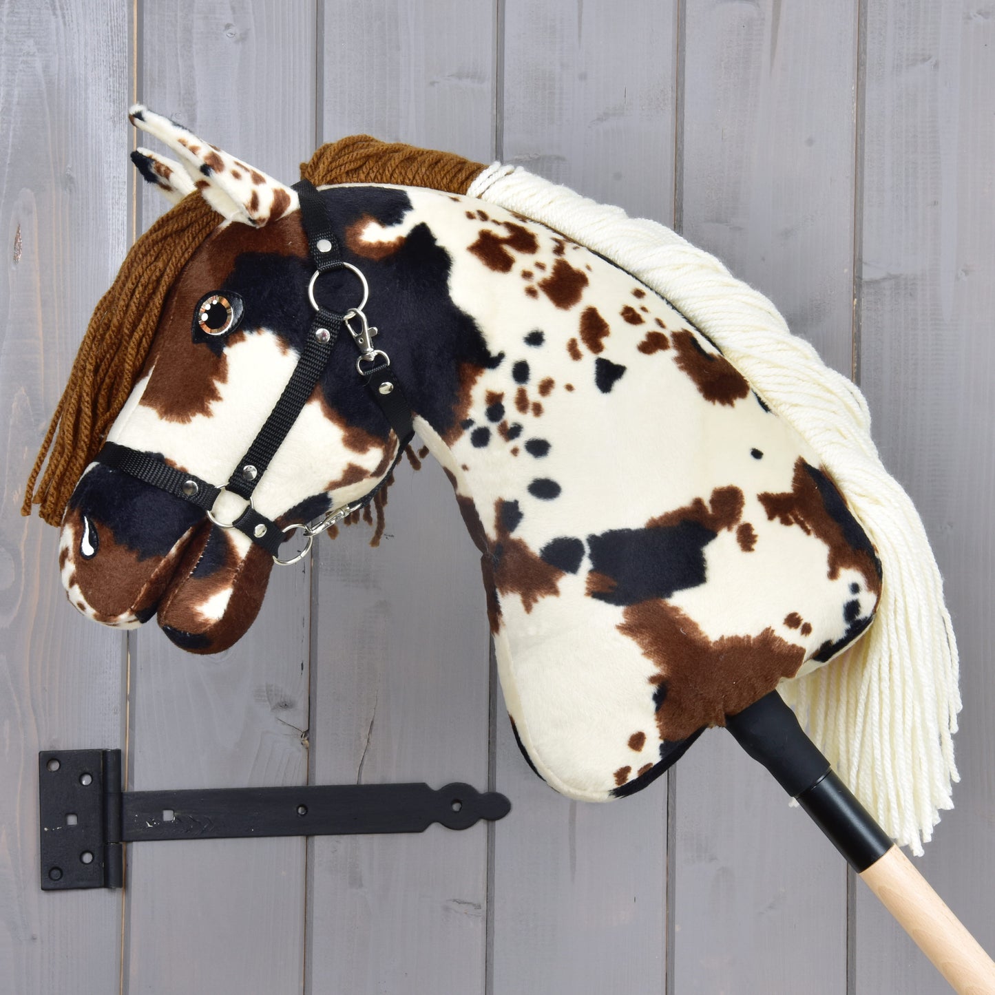 The Hobby Horse Dakota with Black Halter by Hobby Horse LarDen features brown, black, and white spots, a brown yarn mane, and a black halter. Mounted on a wooden stick against a gray wall, its design is suitable for any gender.