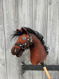 Hobby Horse LarDen's "Hobby Horse Benny with Black Halter" (size XS), styled like a Belgian Warmblood, features a brown body, black mane, and silver details on a wooden stick, shown against a weathered wooden wall.