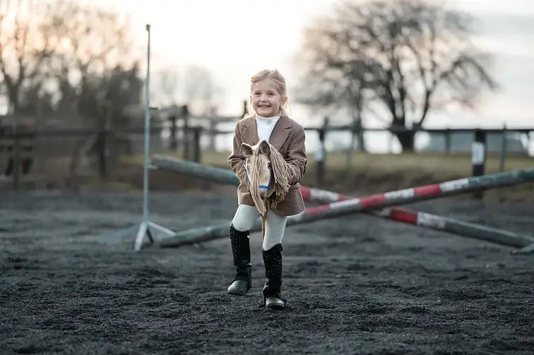 A young girl smiles while enjoying hobby horsing, pretending to ride a toy hobby horse outdoors on a dirt arena with crossed poles and trees in the background. She wears a brown jacket, white pants, and black riding boots.