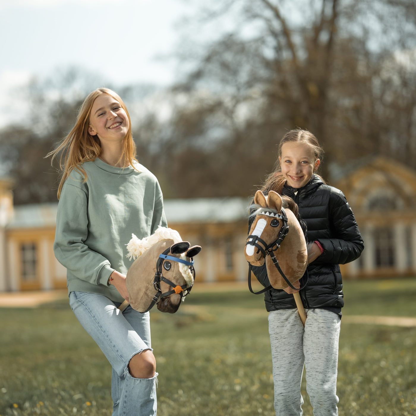 Two smiling girls stand outside holding their hobby horses. One wears a light green sweater and ripped jeans, the other a black jacket and gray pants. Hobby horsing brings them joy as they pose near a large building and trees in the background.