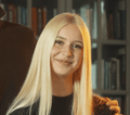 A young woman with long, straight blonde hair smiles at the camera. She is wearing a black top, with a blurred bookshelf behind her. A hand rests gently on her shoulder, suggesting a warm moment after a fun day of hobby horsing.