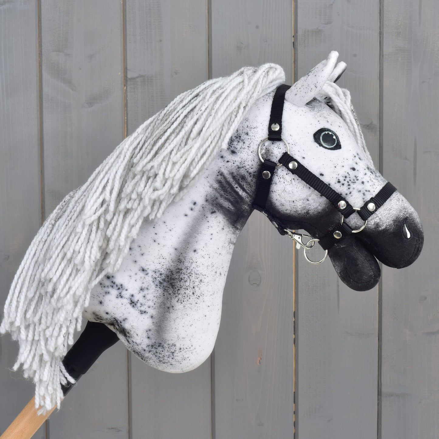 Discover the Hobby Horse LarDen "Hobby Horse Naivy with Black Halter" (size S): a handmade Appaloosa hobby horse, complete with a white and gray yarn mane, bold black spots, and a sleek black halter—set against a rustic wooden backdrop. Perfect for imaginative stallion adventures!.