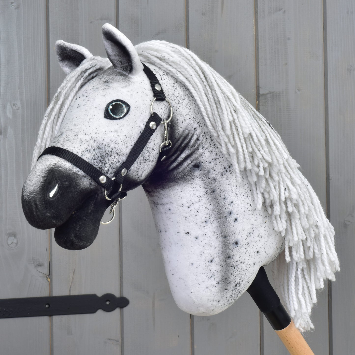 The Hobby Horse LarDen "Hobby Horse Naivy with Black Halter" (size S), featuring a gray and white Appaloosa design with a black halter and yarn mane, is mounted on a wooden stick and set against a gray wooden wall.