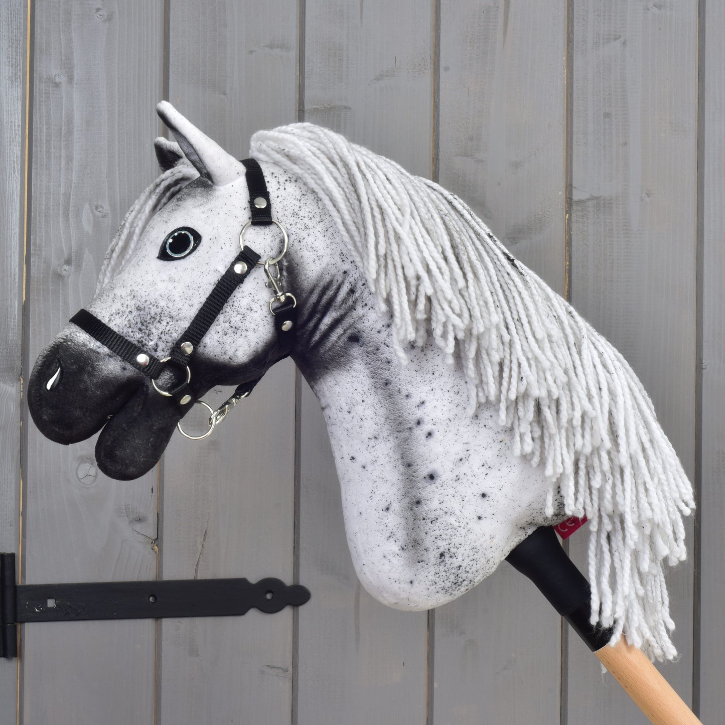 A white and gray Hobby Horse Naivy with a black halter by Hobby Horse LarDen (size S), featuring a yarn mane, is mounted on a wooden stick and positioned against a gray wooden wall.