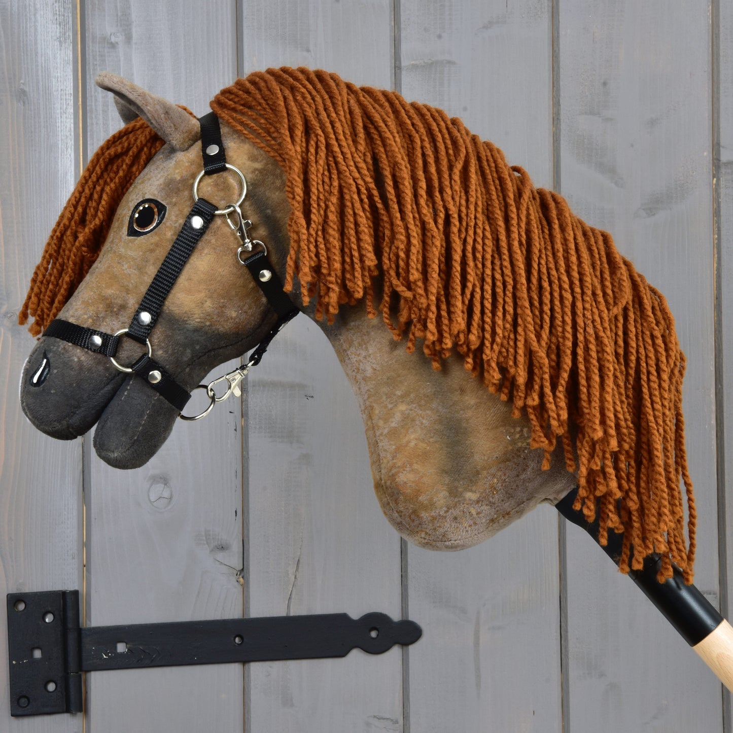 The Hobby Horse LarDen Rafaello with Black Halter (size S), a brown Hanoverian hobby horse with a long yarn mane, is mounted on a wooden wall using a metal bracket.
