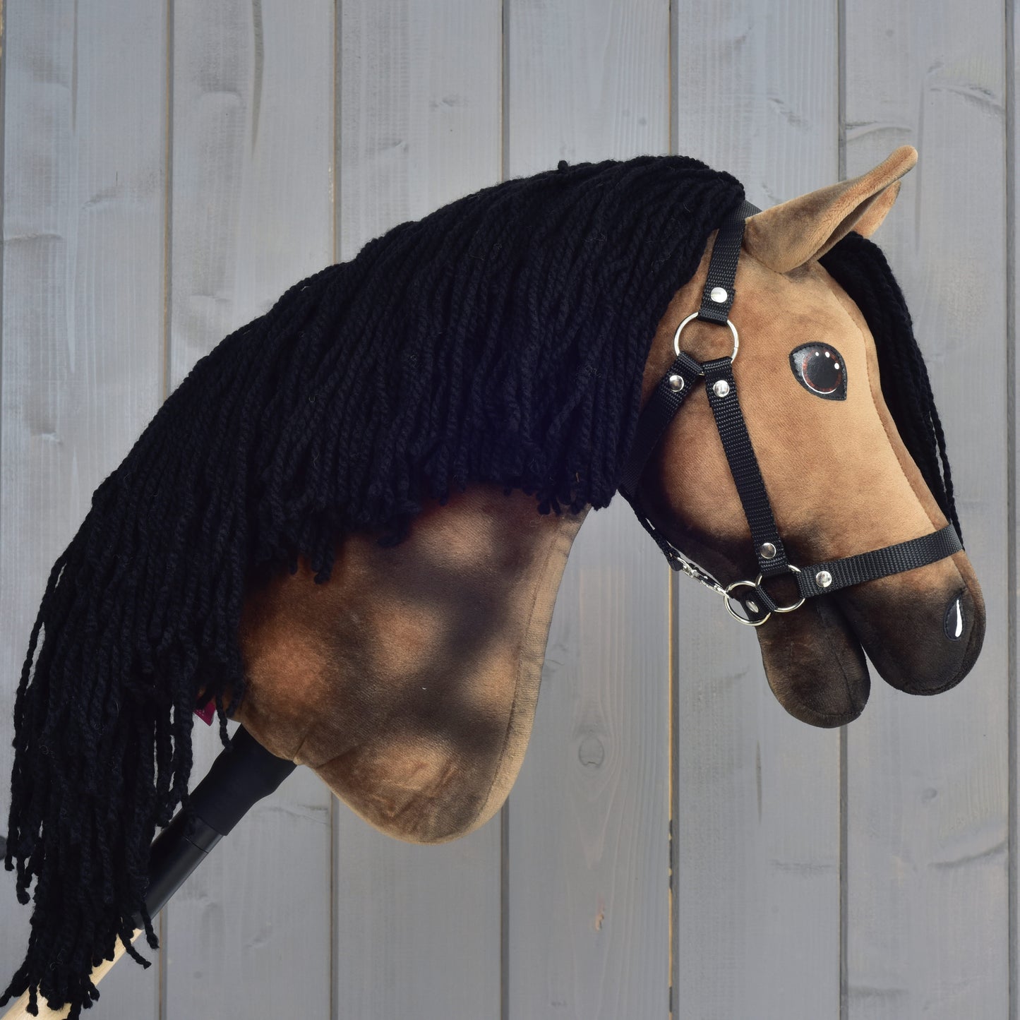 A plush Hanoverian Hobby Horse Stormy with a brown face, black yarn mane, and a black halter from Hobby Horse LarDen is positioned in front of a gray wooden wall.
