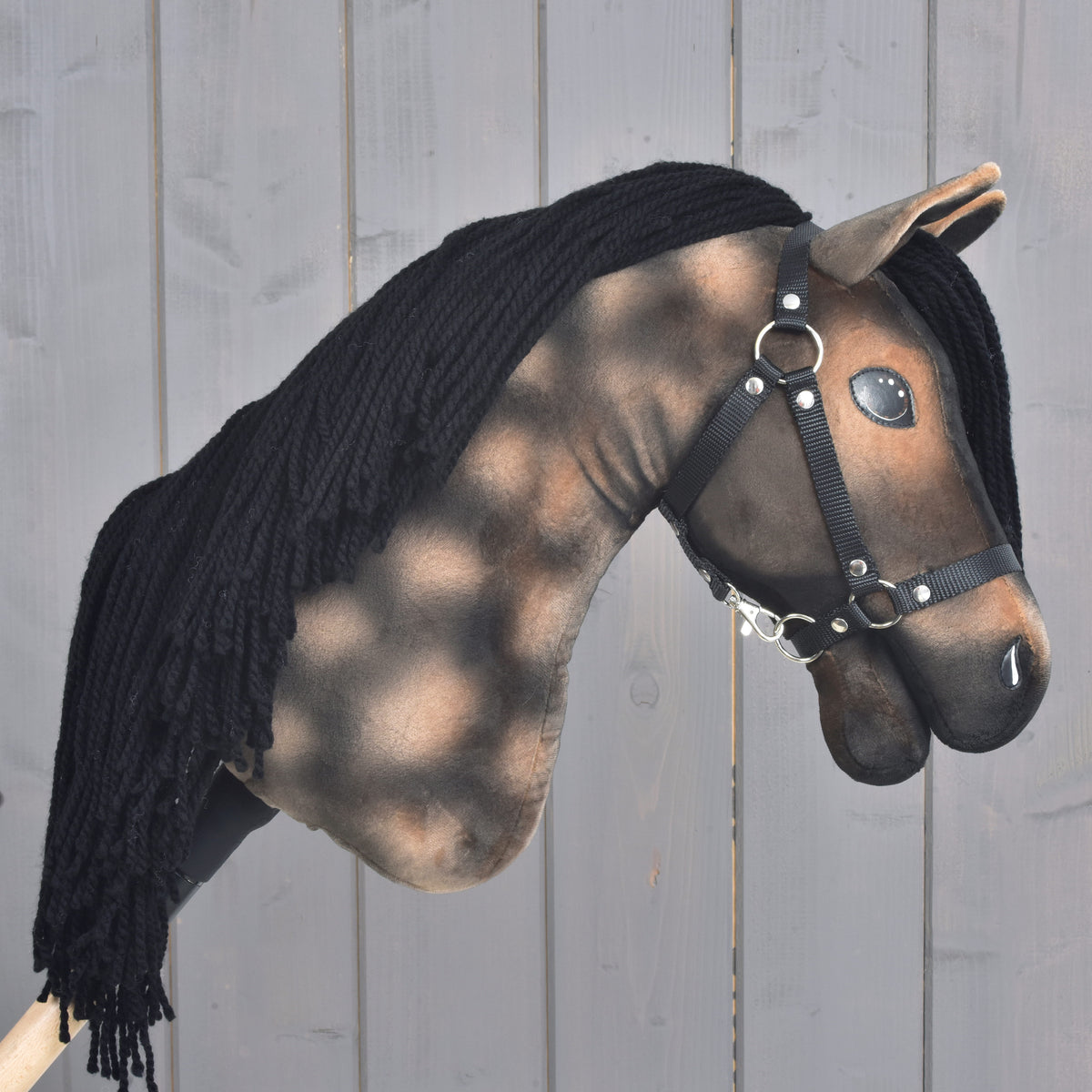 Introducing the Hobby Horse LarDen “Hobby Horse Stormy with Black Halter” — a handcrafted hobby horse inspired by the Hanoverian stallion, featuring a brown and black painted face, black yarn mane, and a stylish black halter, beautifully displayed against a gray wooden background.
