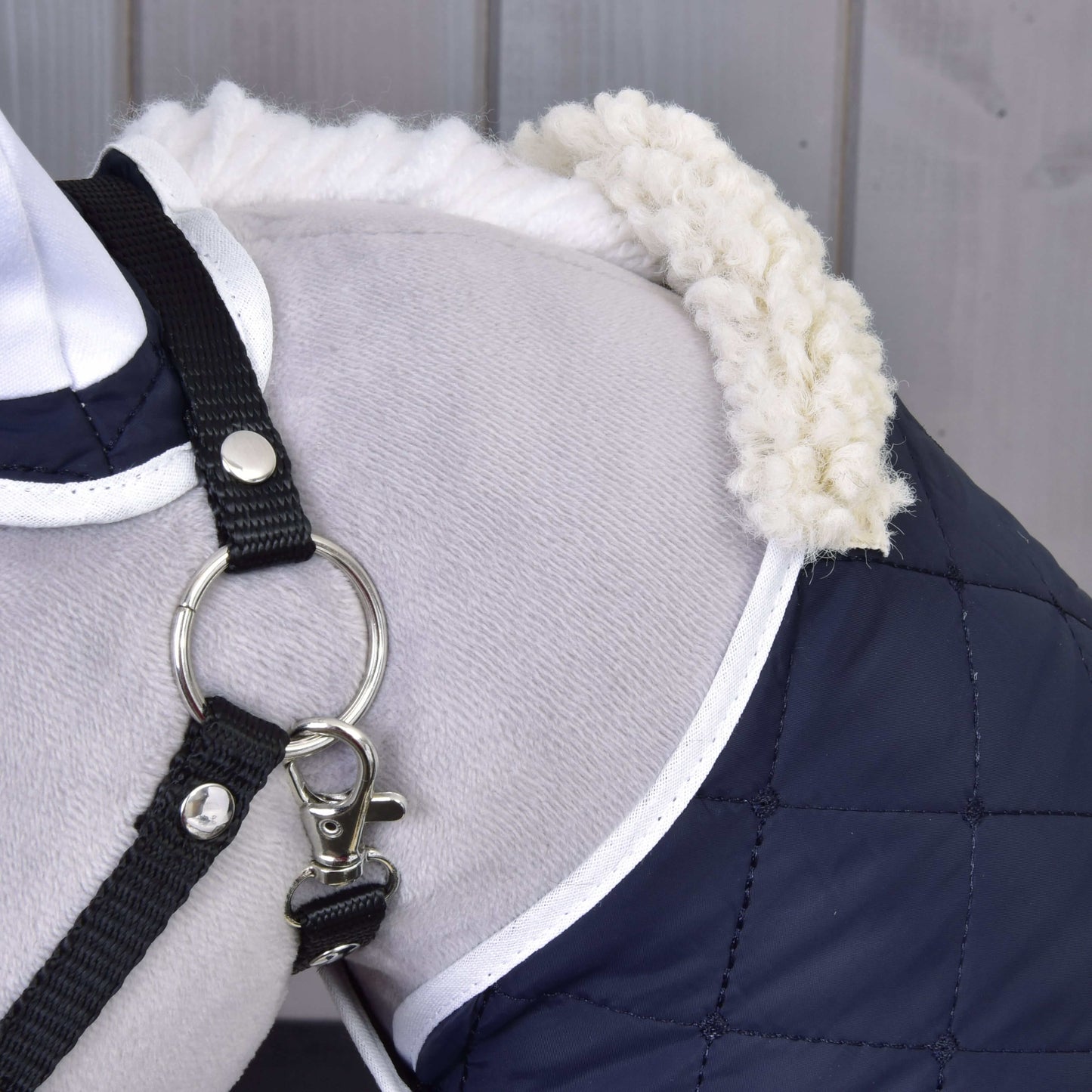 Close-up of a dog's shoulder wearing the Set Thermo Blue coat by Hobby Horse LarDen, featuring quilted navy fabric, white trim and fluffy collar, plus a black harness with metal ring. Wooden paneling is seen in the background.