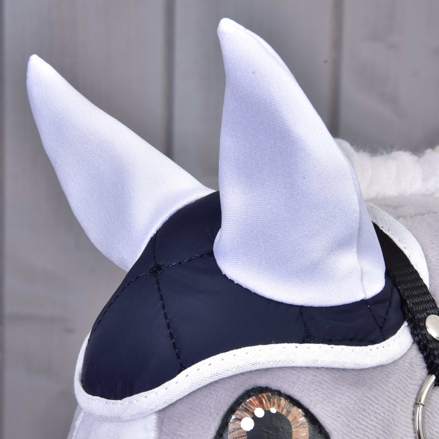 Close-up of a plush horse's head wearing the elegant Set Thermo Blue by Hobby Horse LarDen, featuring white ears, a navy and white cap, and black bridle. The perfect decorative piece against light wooden panels.