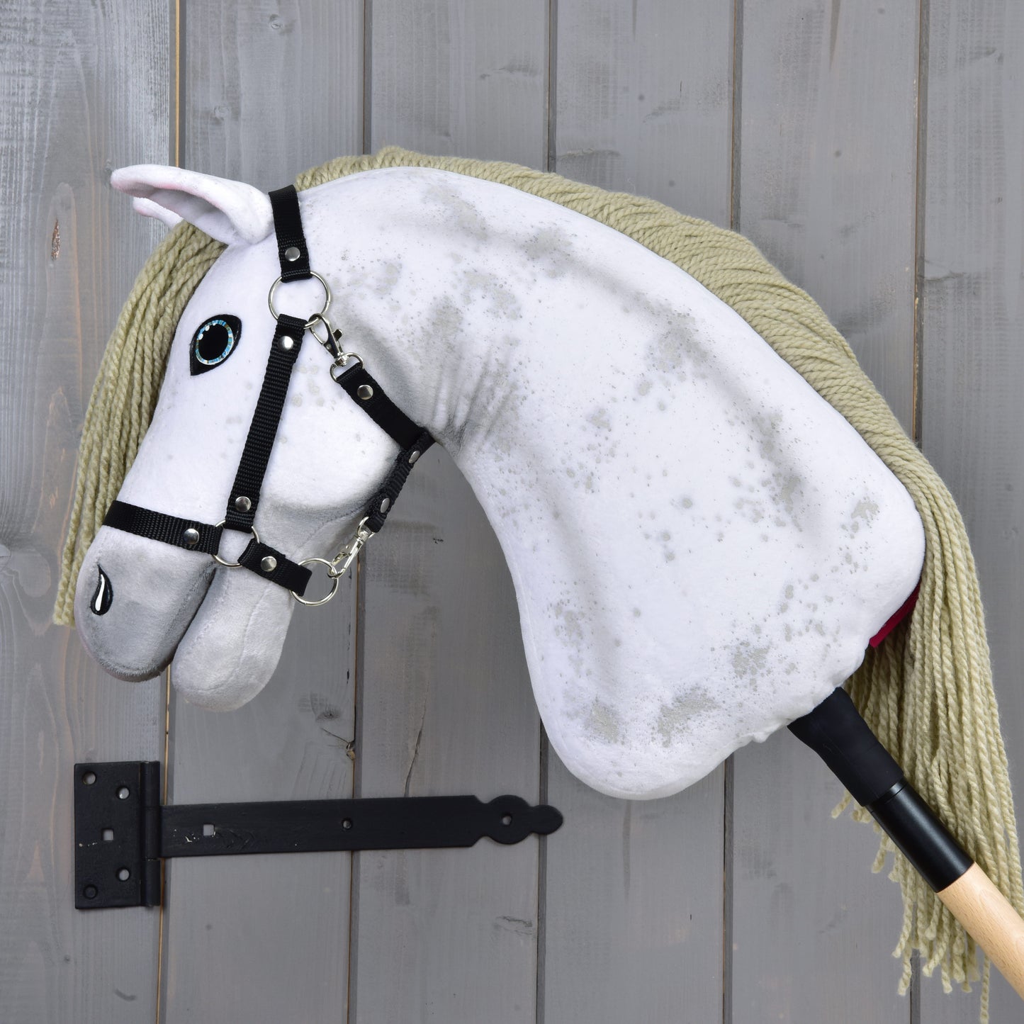 The Hobby Horse Victory with Black Halter (size M) from Hobby Horse LarDen features a white plush design, long yarn mane, and black halter, and is displayed on a gray wooden wall with a black metal bracket.