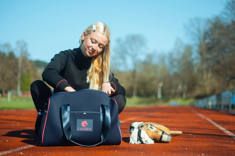 A young woman with long blonde hair sits on a track, looking into a navy sports bag labeled LarDen. A hobby horse lies on the ground beside her, hinting at her passion for hobby horsing. Trees and track lines are visible in the background.