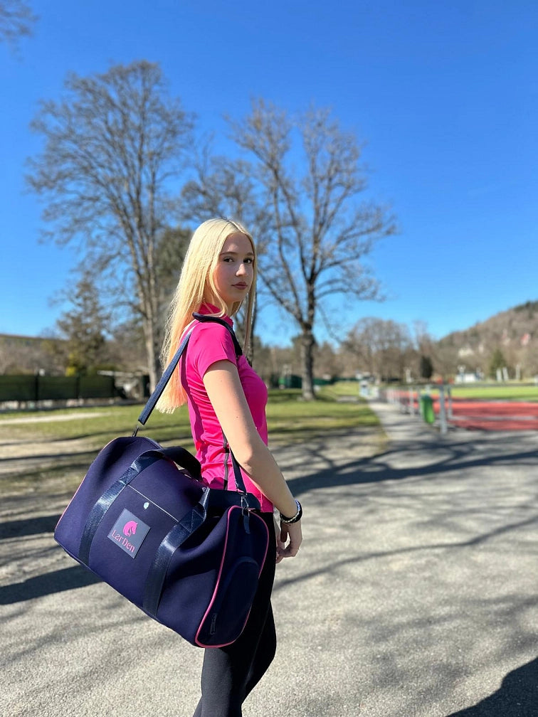 A young woman with long blonde hair, wearing a pink athletic shirt and carrying a navy blue sports bag, stands outdoors on a sunny day with her hobby horse, ready for a fun day of hobby horsing beneath the clear blue sky and leafy trees.