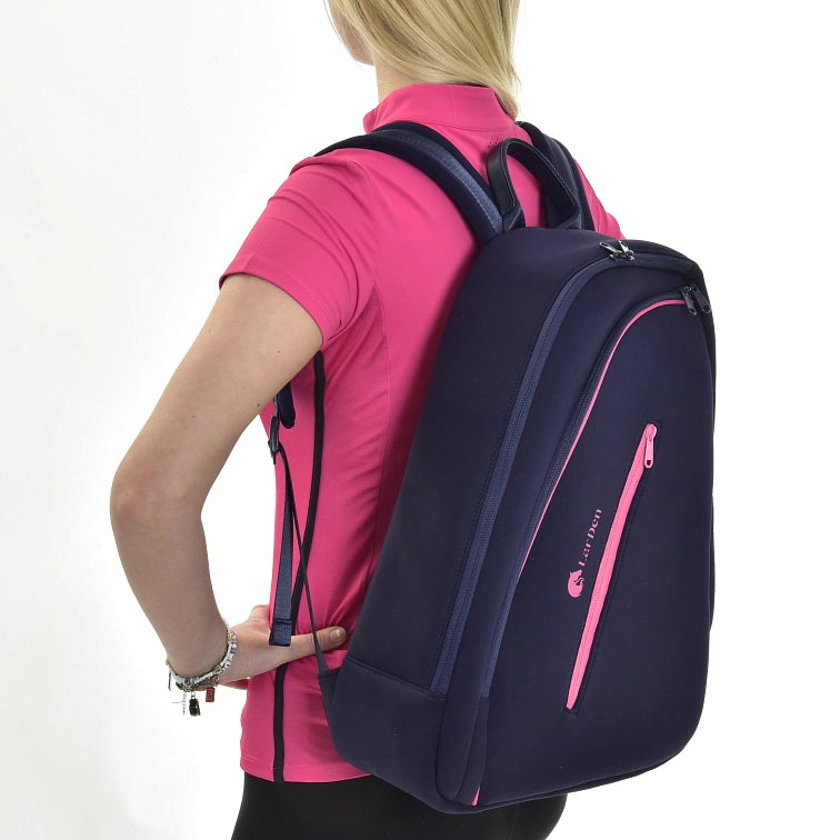 A person with blonde hair wearing a bright pink shirt carries a large navy blue backpack with pink zippers and accents, holding a hobby horse, against a plain white background.