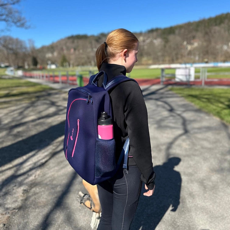 A young woman with red hair in a ponytail walks outside on a sunny day, wearing black athletic clothes and a navy backpack with a pink water bottle. Trees and a track field hint she's heading to or from her favorite Hobby Horsing activity.