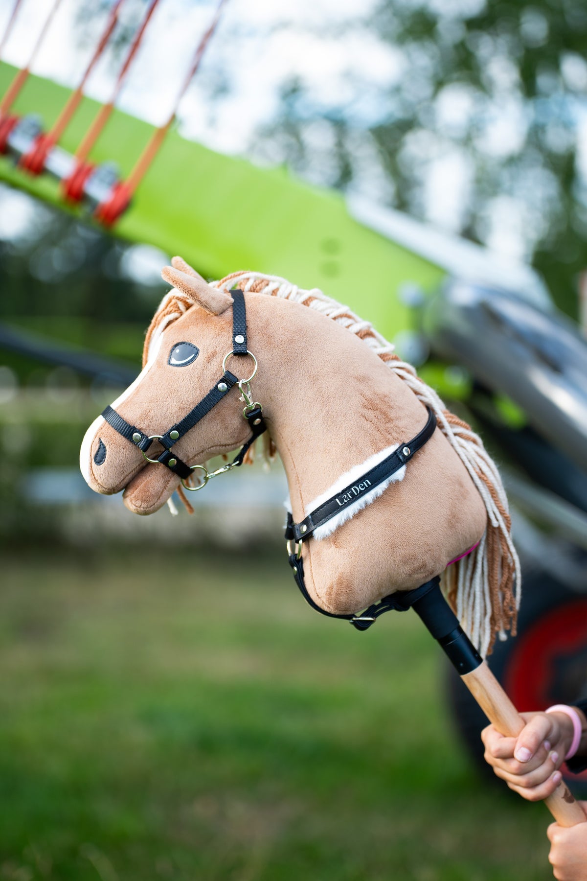 Detail photo: the head and neck of the Hobby Horse Bren with Black Halter by Hobby Horse LarDen in front of a green hay rake - sandy-brown plush, cream yarn mane and forelock - shown here with the Espresso breastgirth with fur.