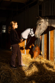 Lifestyle photo: a dark-haired girl in a black sweatshirt stands in the stable aisle holding the Hobby Horse Caesar with Black Halter by Hobby Horse LarDen - grey plush head with a black and white yarn mane - while a grey horse and a bay horse look out of their boxes, shown here with the Complete bitless bridle, decorated.