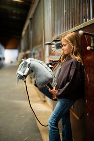 Lifestyle photo: a girl in a dark brown sweatshirt leans against the stalls in a stable aisle holding the Hobby Horse Calypso with Black Halter by Hobby Horse LarDen - grey plush head, pale grey yarn mane - shown here with the fur-lined Teddy Classic halter and the black lead rope.