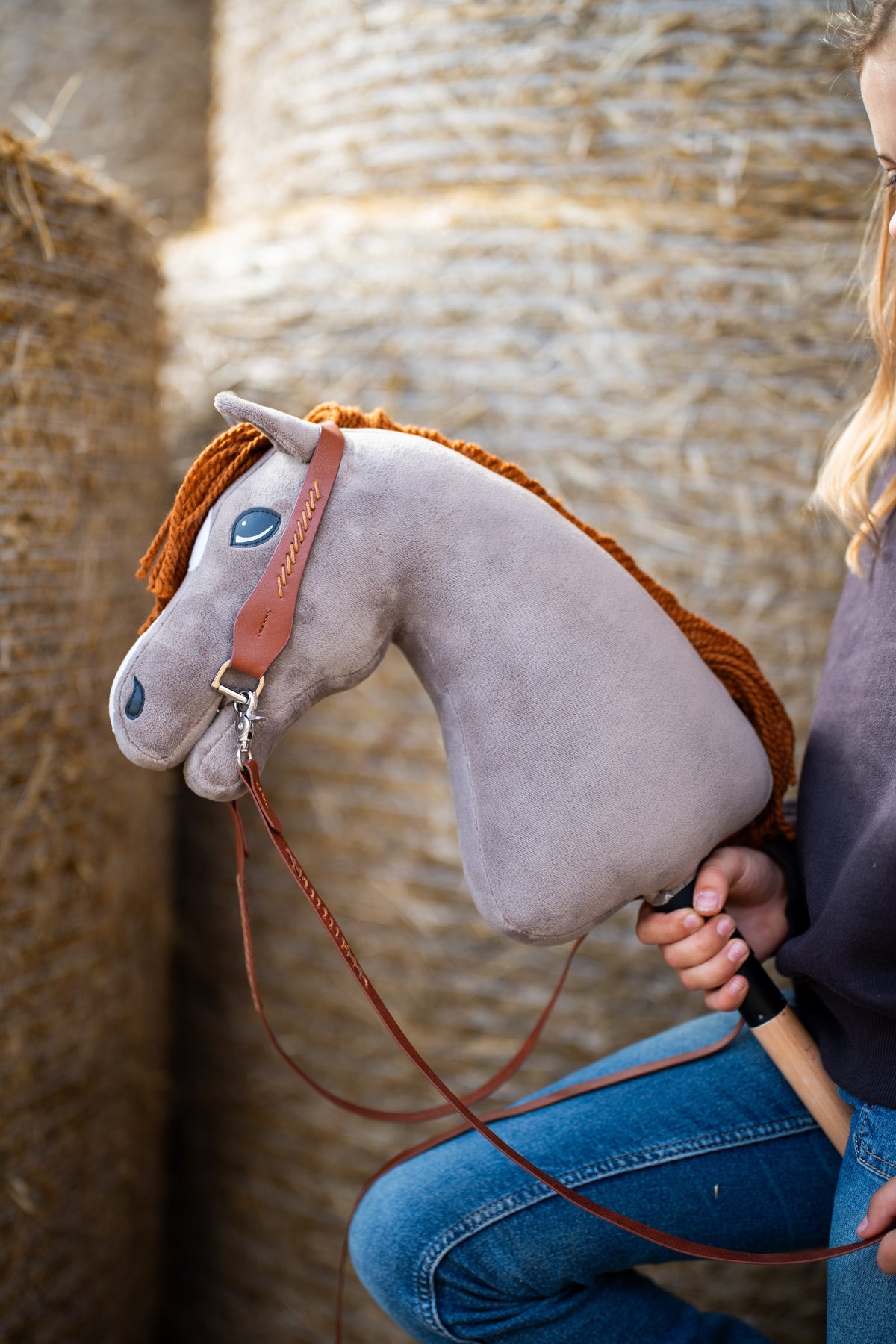 Brown one-ear bridle with reins on a grey plush Hobby Horse with ginger yarn mane beside straw bales