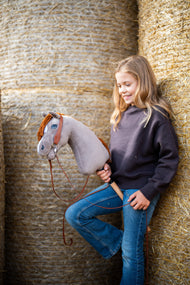 Lifestyle photo: a smiling girl in a dark brown sweatshirt leans against round straw bales holding the Hobby Horse Chanell with Black Halter by Hobby Horse LarDen - grey plush head, blue eye, ginger yarn mane - shown here with the complete Western one-ear bridle.