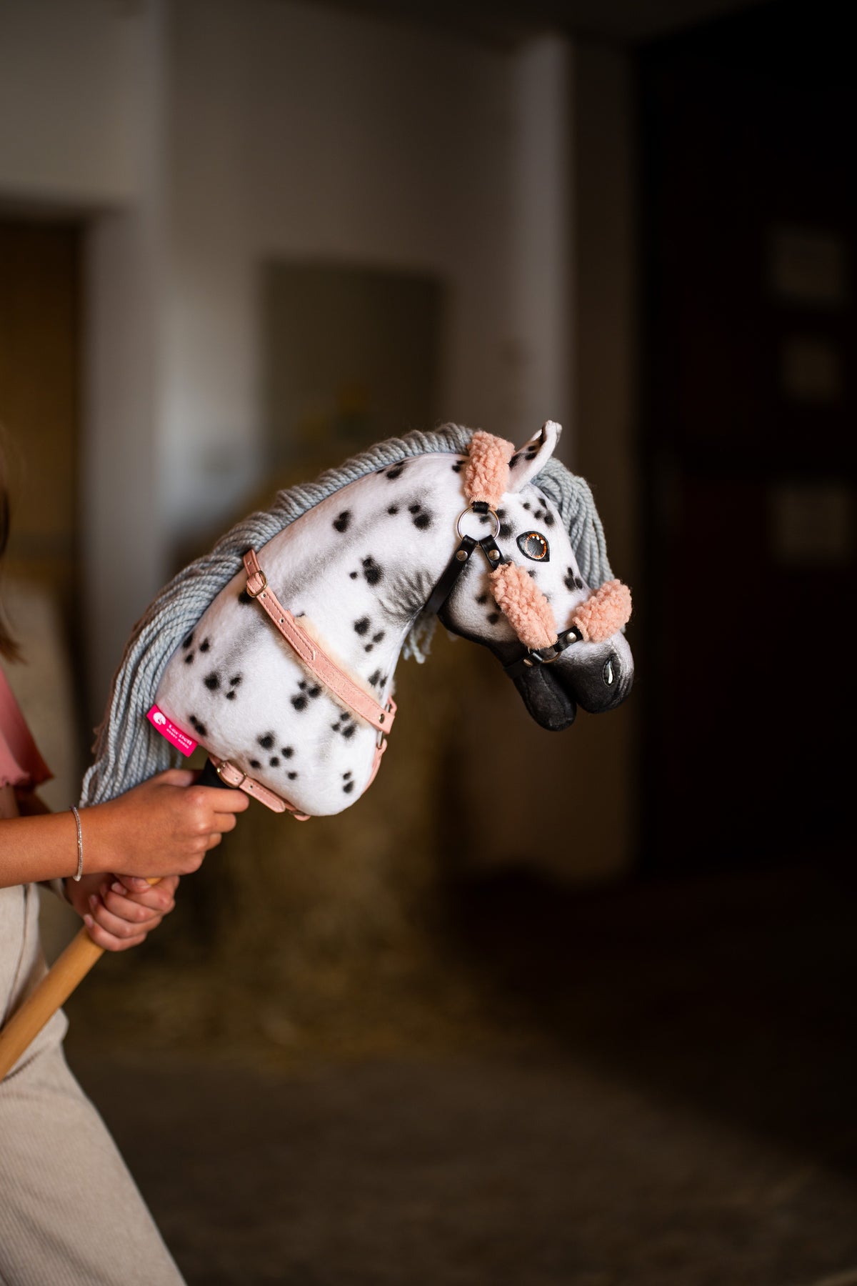 Detail photo: the head and neck of the Hobby Horse Chantal with Black Halter by Hobby Horse LarDen held up inside a stable - white plush with black leopard spots, grey yarn mane, black muzzle - shown here with the Old Rose fur halter in leatherette and the breastgirth with fur.