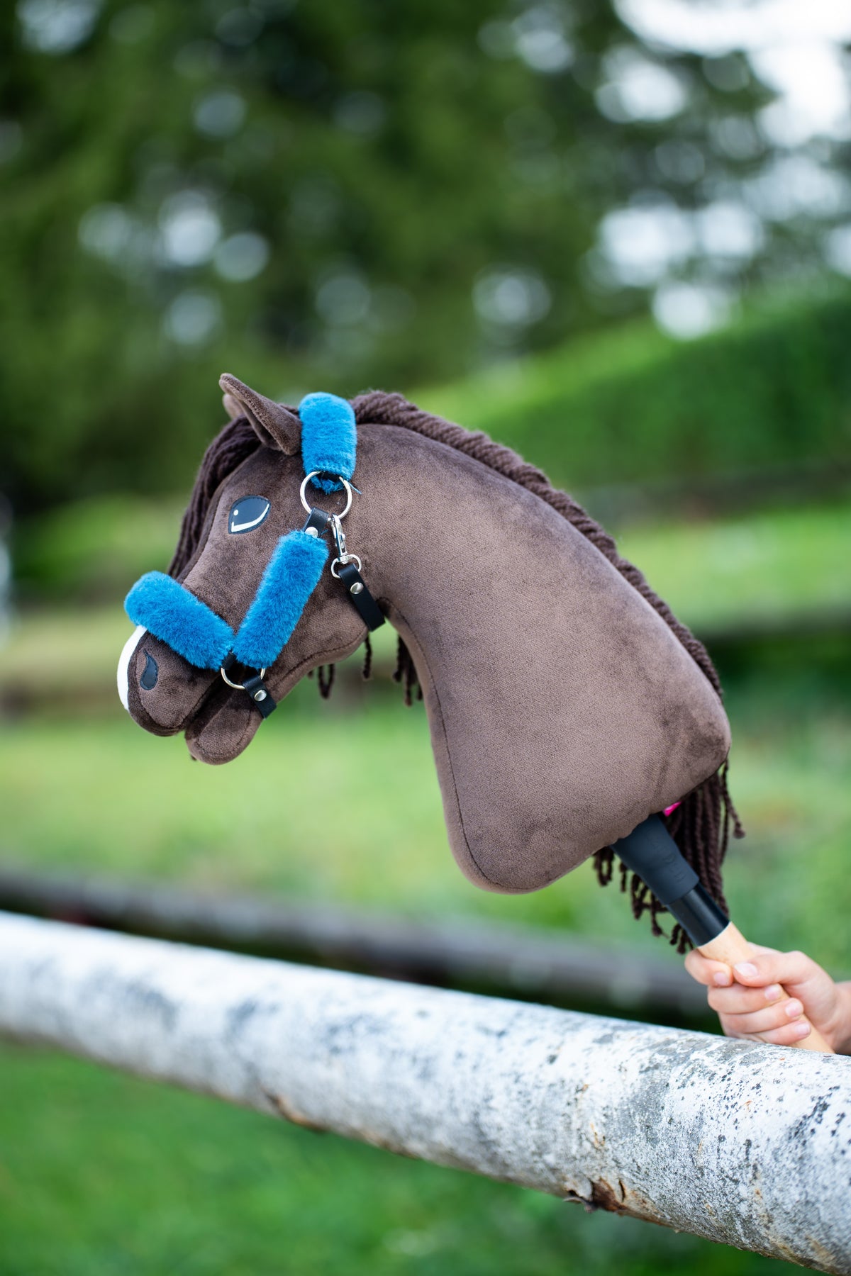 Detail photo: the head of the Hobby Horse Chianti with Black Halter by Hobby Horse LarDen resting over a white paddock rail - dark brown plush, dark yarn mane and forelock - shown here with the fur-lined Teddy Petrol halter in bright blue.