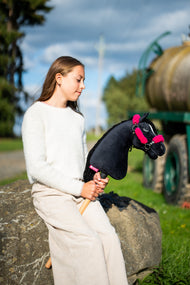 Lifestyle photo: a girl in a cream jumper sits on a boulder by a meadow track holding the Hobby Horse Cloony with Black Halter by Hobby Horse LarDen - black plush head, white star, black yarn mane - shown here with the black leatherette halter with complete pink fur.