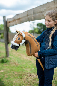 Lifestyle photo: a girl in a navy gilet stands by a wooden paddock fence holding the Hobby Horse Connie with Black Halter by Hobby Horse LarDen - chestnut plush head with a dark dorsal stripe, cream yarn mane - shown here with the fur-lined Teddy Classic halter.