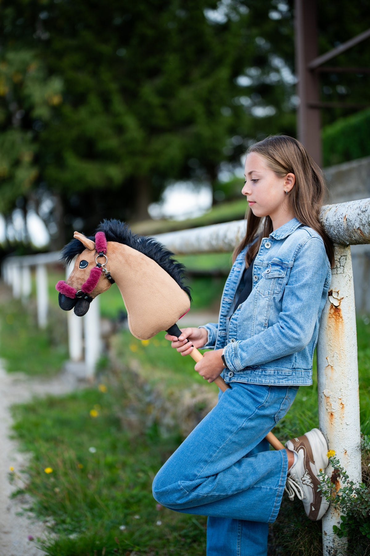 Lifestyle photo: a girl in double denim leans against a white paddock rail holding the Hobby Horse Cookie with Black Halter by Hobby Horse LarDen - sandy plush head, black yarn mane - shown here with the fur-lined Teddy Maroon halter.