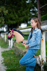 Lifestyle photo: a girl in double denim leans against a white paddock rail holding the Hobby Horse Cookie with Black Halter by Hobby Horse LarDen - sandy plush head, black yarn mane - shown here with the fur-lined Teddy Maroon halter.