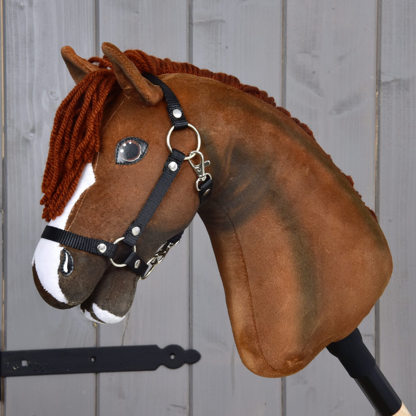 The Hobby Horse LarDen "Hobby Horse Cooper with Black Halter," a brown plush hobby horse featuring white markings and a black halter, is mounted on a stick and set against a gray wooden background.