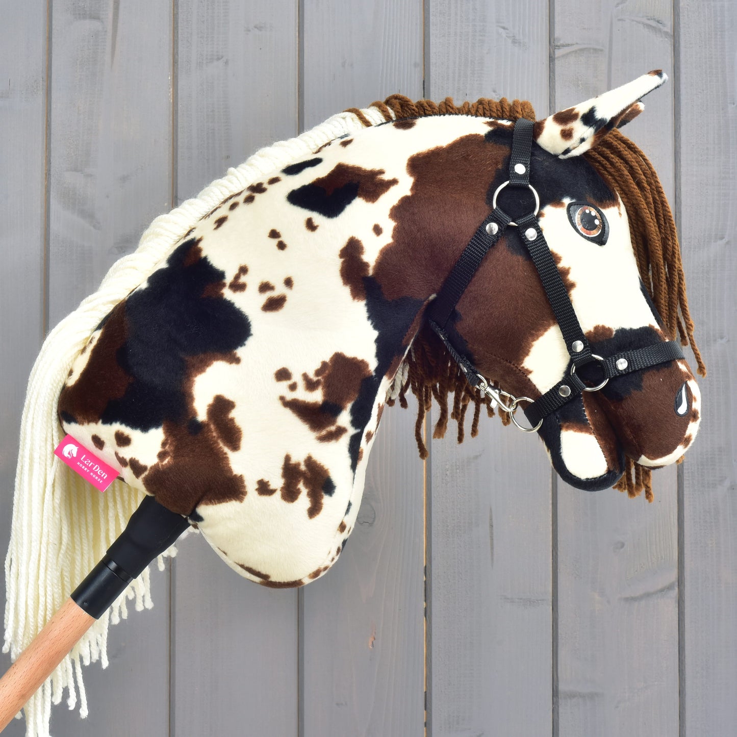 The Hobby Horse Dakota with Black Halter by Hobby Horse LarDen captures the untamed spirit of the Mustang with its brown and white spotted plush design, black halter, and yarn mane. Mounted on a wooden stick, it's displayed against a light wooden plank background evocative of North America's open plains.