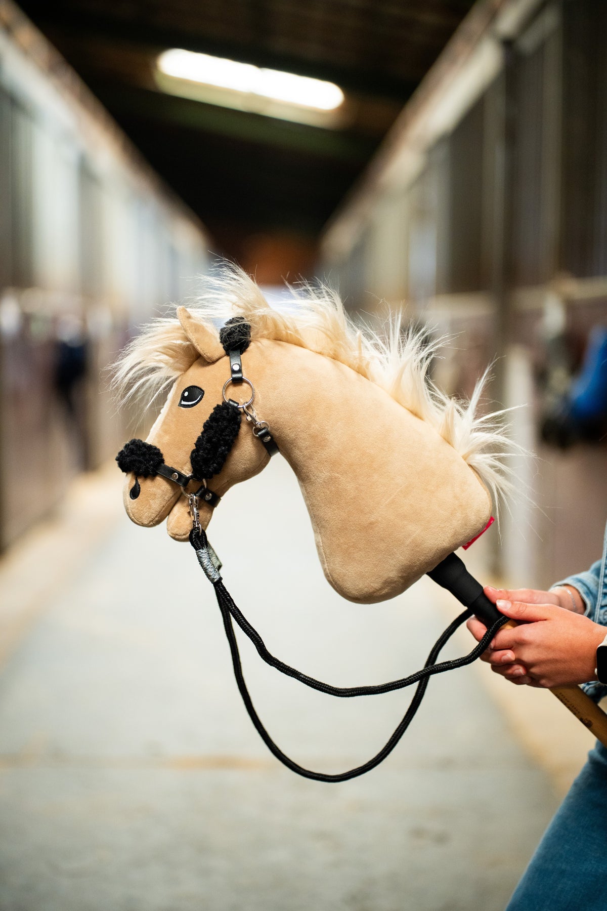 Detail photo: the head of the Hobby Horse Dancing Lady with Black Halter by Hobby Horse LarDen held up in a stable aisle, the reins looping down - cream plush, pale yarn mane - shown here with the black leatherette halter with complete black fur and the black reins.