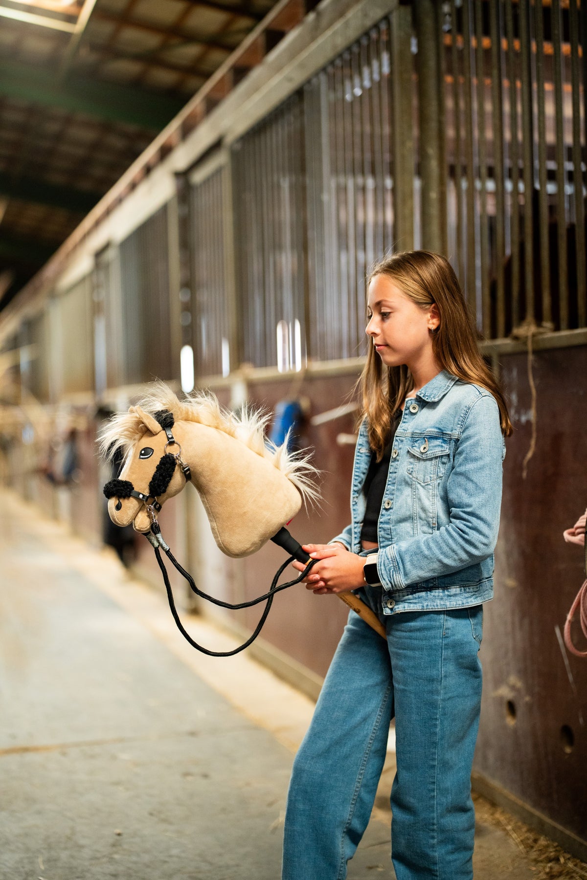 Lifestyle photo: a girl in a denim jacket walks the Hobby Horse Dancing Lady with Black Halter by Hobby Horse LarDen down a stable aisle - cream plush head, pale yarn mane and forelock - shown here with the black leatherette halter with complete black fur and the black reins.