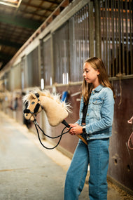 Lifestyle photo: a girl in a denim jacket walks the Hobby Horse Dancing Lady with Black Halter by Hobby Horse LarDen down a stable aisle - cream plush head, pale yarn mane and forelock - shown here with the black leatherette halter with complete black fur and the black reins.