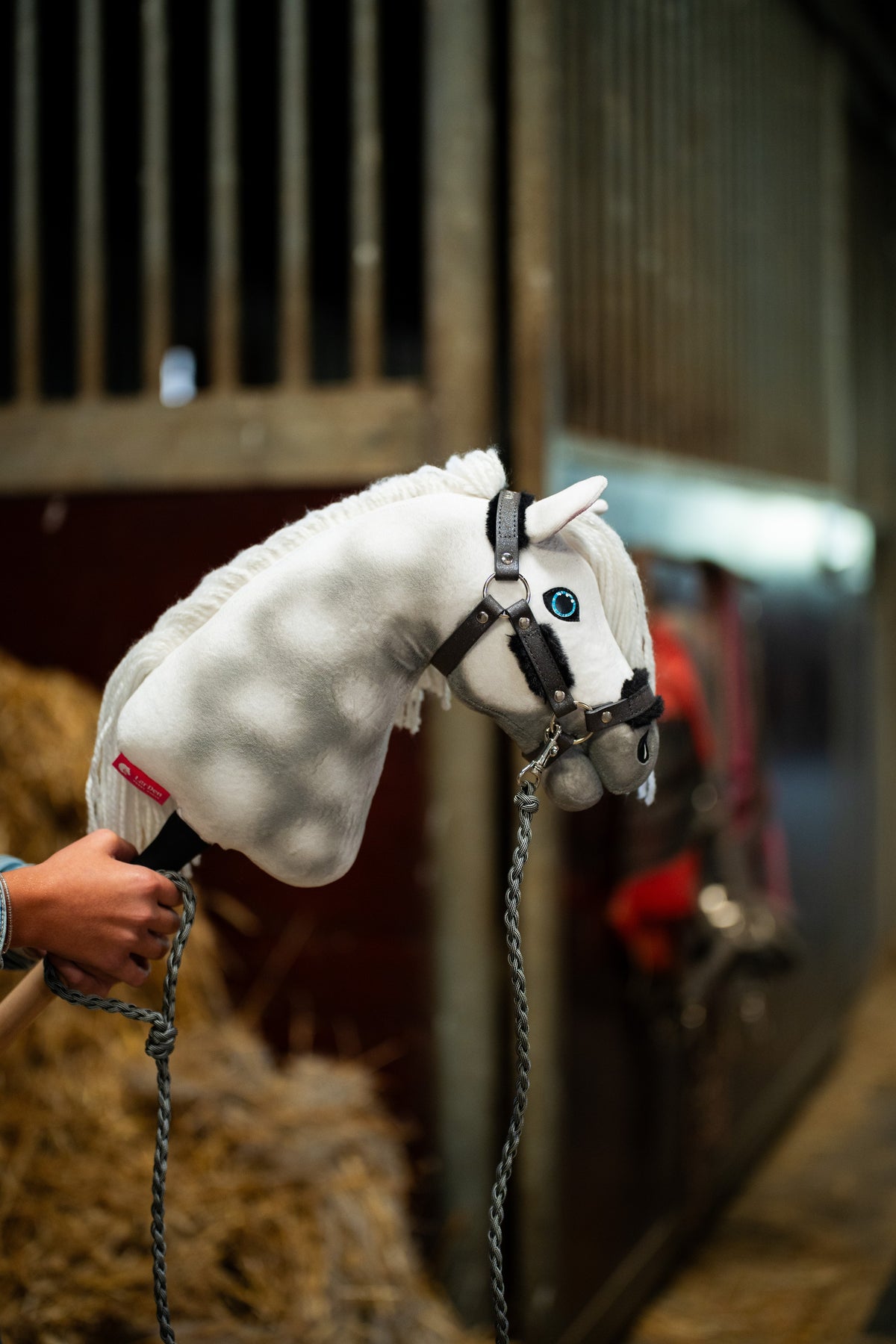 Detail photo: the head of the Hobby Horse Elektra with Black Halter by Hobby Horse LarDen held up by the stalls, a rug-covered horse blurred behind - white dappled plush, blue eye, white yarn mane - shown here with the Graphite Full fur halter in leatherette and the knitted lead rope.