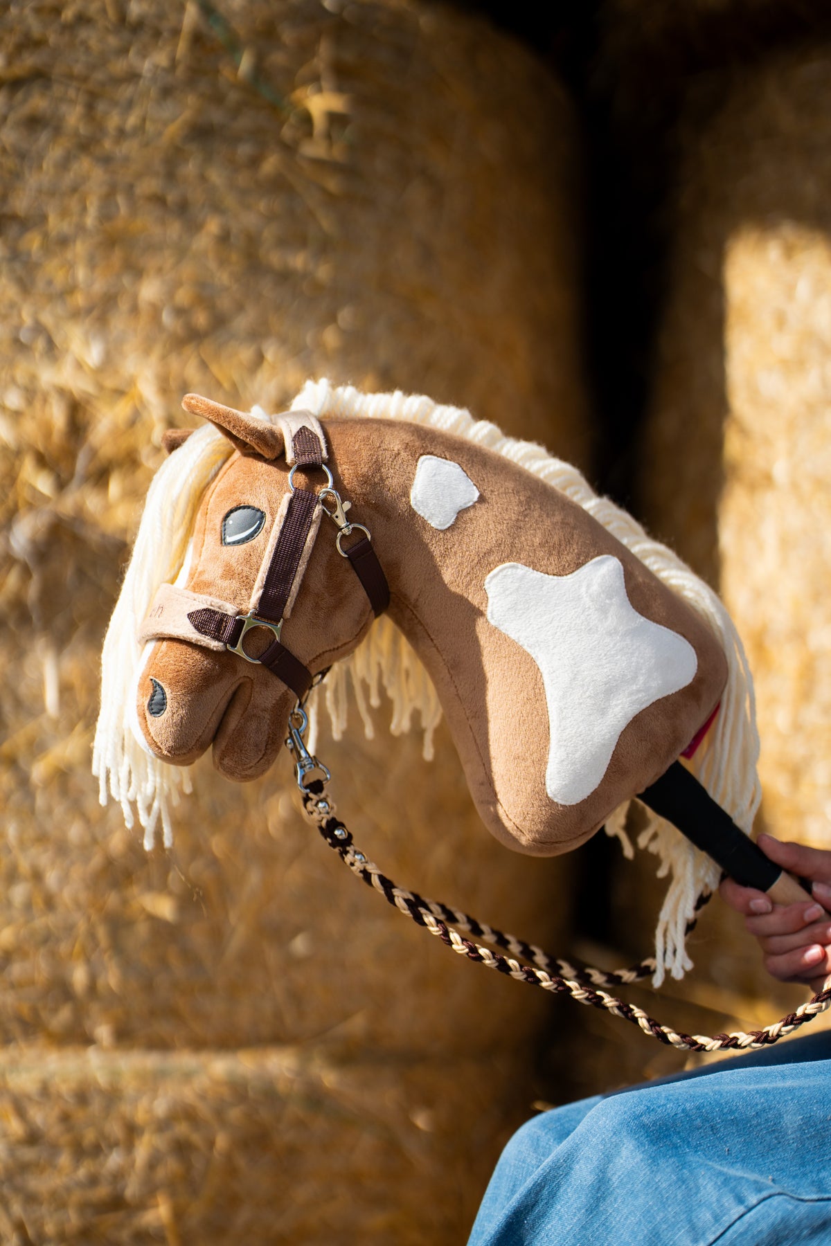 Detail photo: the head and neck of the Hobby Horse Fury with Black Halter by Hobby Horse LarDen in front of round straw bales - palomino plush with white markings, cream yarn mane - shown here with the fur-lined LarDen Brown halter and the braided Bloom Brown lead rope.