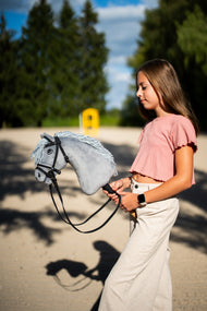 Lifestyle photo: a girl in a pink top walks the Hobby Horse Grace with Black Halter by Hobby Horse LarDen across a sand arena - grey plush head, pale grey yarn mane - shown here with the complete De Luxe bridle with two browbands.