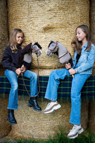 Lifestyle photo: two girls sit on straw bales on a tartan blanket, each holding a Hobby Horse Guccini by Hobby Horse LarDen - grey Holsteiner-style head with a white nose stripe and a long dark brown yarn mane - shown here with the fur-lined Walnut halter, the Walnut breastgirth with fur and the Vivaldi lead rope.