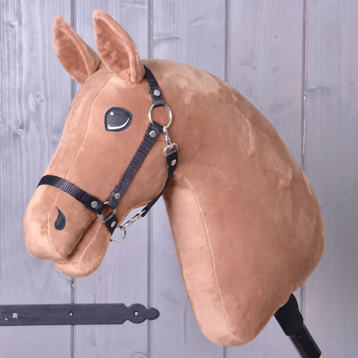 A plush hobby horse head with a bridle, mounted on a stick, rests against a wooden wall background—ideal inspiration for creating your own unique horse using the “Build your horse” configurator from Hobby Horse LarDen before completing your purchase.