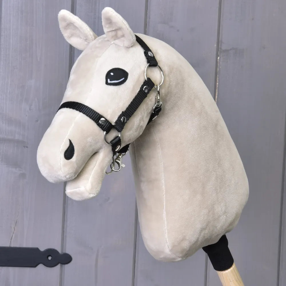 A “Build your horse” plush custom hobby horse by related_to_12441055625556, featuring a light beige color, striking blue eyes, and a black harness attached, is mounted on a wooden stick and positioned against gray wooden paneling.