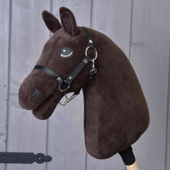 The Build Your Horse plush custom hobby horse head, in dark brown with a black bridle and reins and striking white manes, is attached to a wooden stick and displayed in front of gray wooden panels.