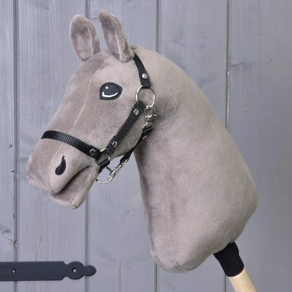 The "Build your horse" custom hobby horse from related_to_12441055625556 features a gray plush head with striking blue eyes and a black bridle, mounted on a wooden stick, and is displayed in front of a gray wooden door.